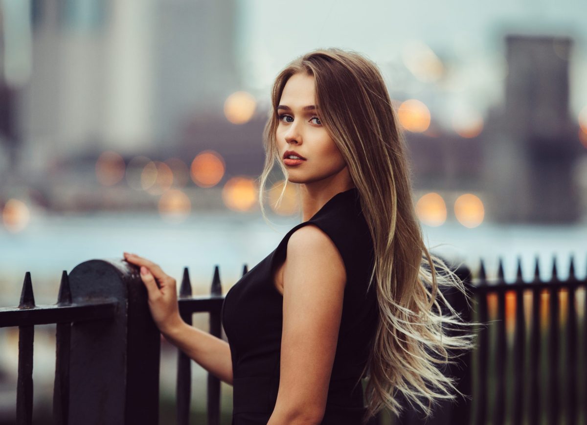 Gorgeous young model woman with perfect blonde hair looking at camera posing in the city wearing black evening dress.